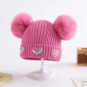 Pink knit baby beanie with double pom-poms and glitter heart decor on white display