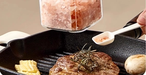 Pink Himalayan salt in a clear jar being sprinkled onto a steak with a small spoon