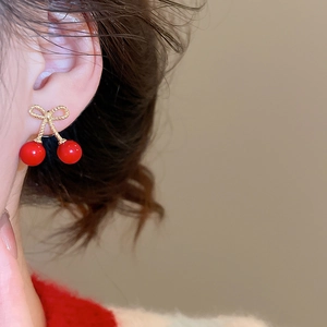 Detail of red cherry stud earrings with golden bow design worn on female model's ear