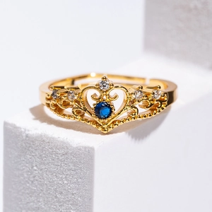 Golden heart crown ring with blue gemstone and clear crystals on a white pedestal