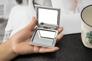 Hand holding an open portable square silver metal compact mirror for makeup touch-ups