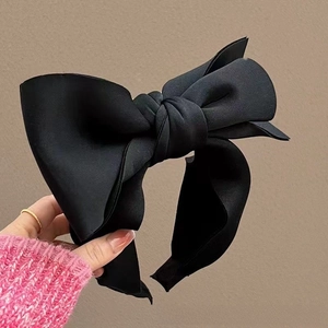 Close-up of a stylish black oversized satin bow headband held against a neutral wall