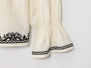 Detail of cream white cotton linen blouse featuring black ethnic embroidery and ruffled cuffs