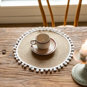Close-up of beige jute placemat with white pom-pom fringe and ceramic teacup on wood table