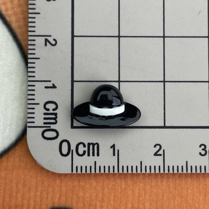 Close-up of a small black resin hat charm measured on a metric ruler for size clarity