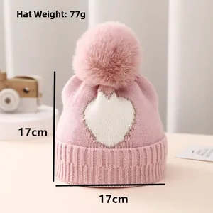 Detailed view of pink knitted baby hat with white heart pattern and large faux fur pompom