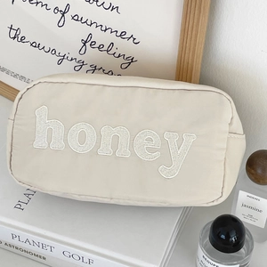Detail view of cream nylon pouch with honey letter embroidery and gold zipper closure