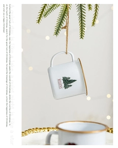 Close-up of white enamel Christmas cup ornament with green tree and gold rim on pine branch