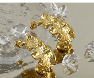 Detailed view of 18K gold plated asymmetric wavy earrings on reflective ice props