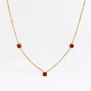 Détail d'un collier en acier doré avec trois pendentifs en zircon rouge carrés sur fond blanc