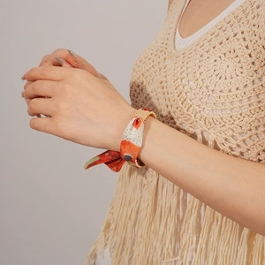 Close-up of matte gold metal buckle and patterned orange silk scarf bracelet on model