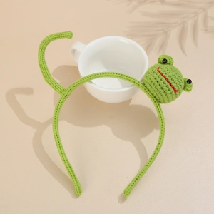 Detailed view of a green hand-knitted frog headband for kids on a ceramic cup base