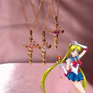 Three golden sword necklaces with winged heart gems in pink and purple on pink backdrop