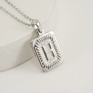 Macro shot of silver rectangle pendant featuring embossed letter H and sunburst texture