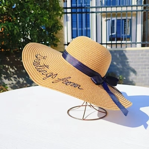 Beige wide brim straw sun hat with navy ribbon and Greetings From embroidery on white table