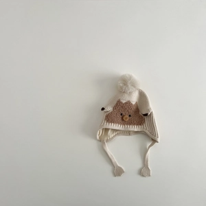 Detailed close-up of cream knit winter earflap hat with brown bear patch and pompom