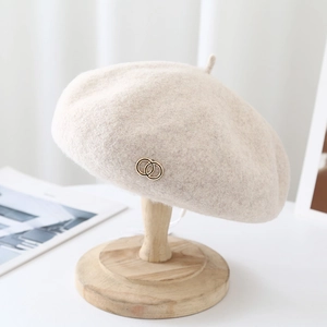 Detail view of cream wool beret with gold double-ring emblem on a wooden stand