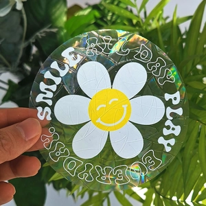 Detailed holographic suncatcher sticker featuring a smiley daisy held against green leaves