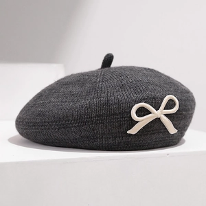 Détail du tricot d'un béret gris foncé avec un nœud crème sur socle blanc épuré