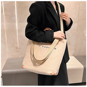 Model wearing a large beige canvas tote bag with brown leather handles and shoulder strap
