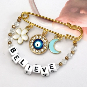 Detail of golden safety pin brooch featuring evil eye charm and BELIEVE letter beads
