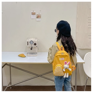 Kid modeling bright yellow backpack with plaid pocket and white bunny toy in cozy cafe