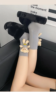 Detail of gray knitted socks with 3D teddy bear charm and plush black slippers on a sofa