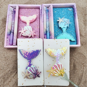 Flat lay of mermaid notebooks with sequins and seashell charms on a sandy beach