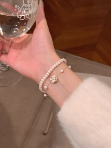 Primer plano de pulsera de doble capa con perlas y colgante de camelia dorada en la muñeca