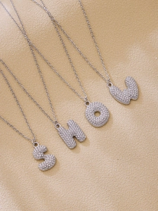 Detailed shot of silver zircon initial necklaces spelling SHOW on textured background