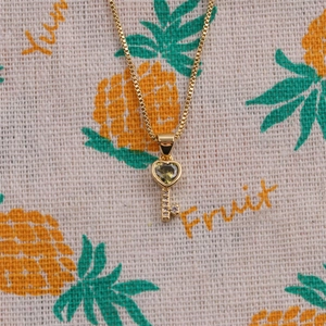 Detailed close-up of a gold-plated key necklace featuring a green heart crystal and micro-pavé