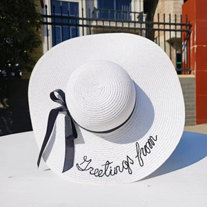 Close-up of a white wide-brim straw hat with black Greetings from embroidery and a bow