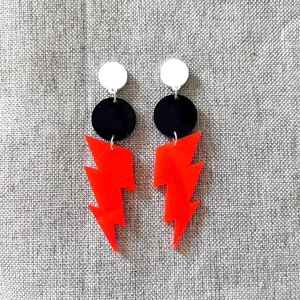 Neon lightning bolt earrings with red acrylic charms and round studs on grey linen texture