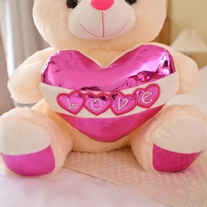 Detailed macro view of a plush beige teddy bear holding a metallic pink LOVE heart pillow