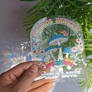 Holographic suncatcher sticker with mushroom and butterfly designs held against sunlight
