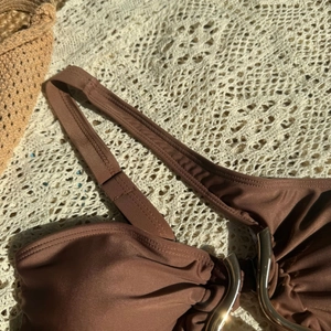 Detail view of a brown bikini top with a golden ring on a woven beach fabric background