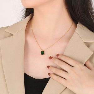 Model wearing a gold necklace with a square green gemstone pendant on a brown blazer