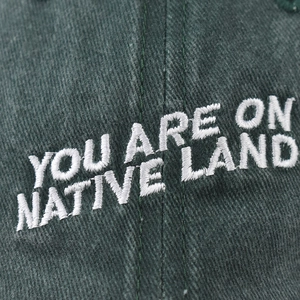 Macro shot of white 'YOU ARE ON NATIVE LAND' embroidery on washed green baseball cap