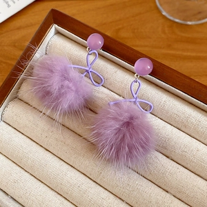Dettaglio di orecchini a pompon in pelliccia viola con fiocchi metallici in portagioie morbido