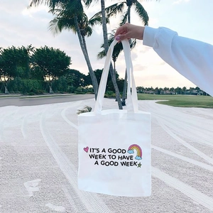 White canvas tote bag with 'It's a good week' print held in a tropical beach setting