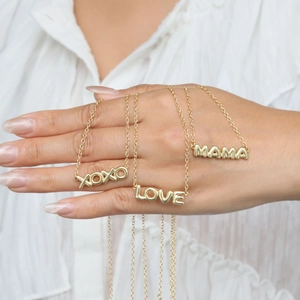 Close-up of gold LOVE, MAMA and XOXO alphabet pendant necklaces held by a model