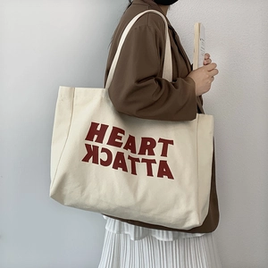 Model wearing a large beige canvas tote bag featuring a red Heart Attack letter print