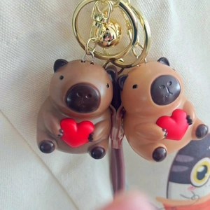 Miniature capybara keychain charms holding red hearts with golden bells and hooks
