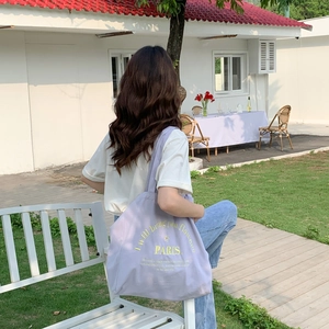 Back view of a woman carrying a lavender canvas tote bag with yellow embroidery in a garden