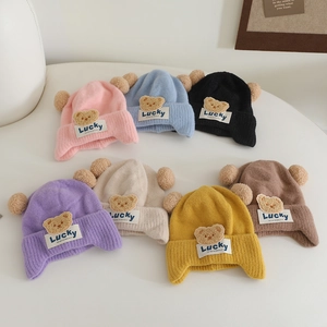 Detailed flat lay of children's knit beanies in seven colors with dual pompoms and bear patch