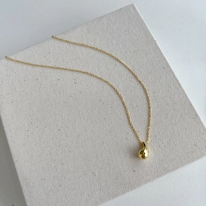 Detail of a polished golden bean pendant on a minimalist thin chain displayed on white stand