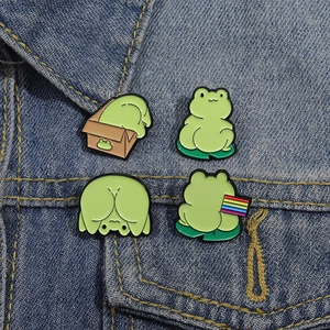 Set of four cute cartoon green frog enamel pins displayed on a blue denim jacket pocket