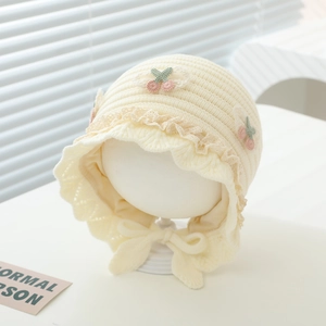 Detail of baby knit bonnet with lace trim, cream cotton yarn, and crochet cherry applique