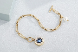 Detail of gold paperclip bracelet with blue gemstone pendant and pearl charm toggle clasp