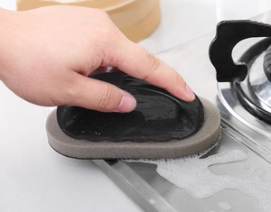 Close-up of handheld emery sponge brush cleaning stains on a kitchen stovetop surface